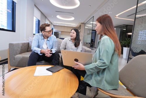 Asian female marketing and sales staff holding laptop explaining products to Asian male and female staff businessmen colleagues sitting in living room in company office and having discussion negotiati