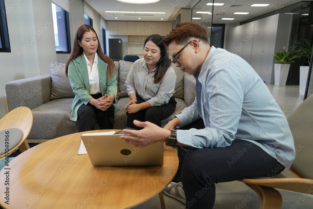 An Asian male sales and marketing representative sits explaining information to two Asian female customers with a laptop in the company's living room.