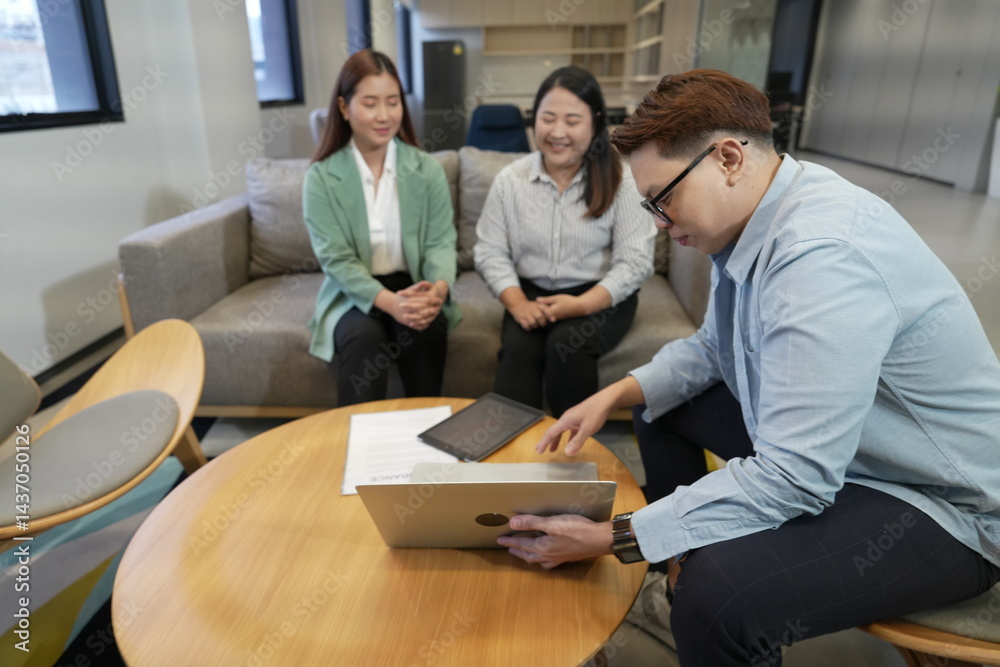 An Asian male sales and marketing representative sits explaining information to two Asian female customers with a laptop in the company's living room.