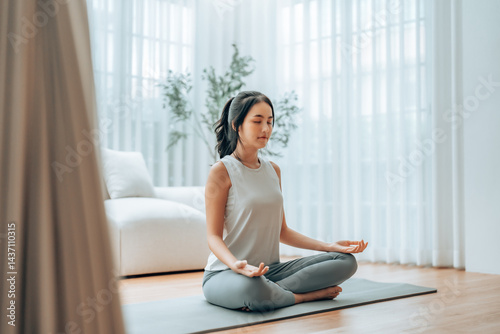 Young Asian woman meditating in lotus position at home, sitting on the floor in fitness clothing. Breathing workout to achieve relaxation, peace and mindfulness