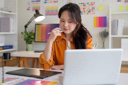 Woman immersed in design thinking, reviewing her work on a laptop in a bright, organized studio with visual inspiration on the walls.