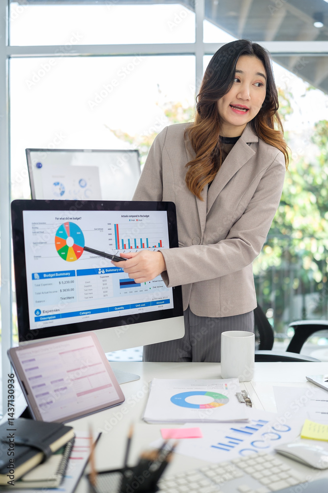 Businesswoman in formal attire explaining budget charts and analytics on a computer screen during a business presentation in a modern office. Businesswoman in formal attire explaining budget charts and analytics on a computer screen during a business presentation in a modern office.