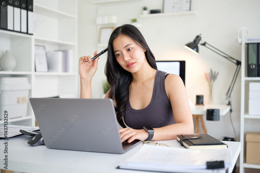 Obraz premium Asian woman sitting at home office desk and focusing on her laptop screen in a moment of contemplation.