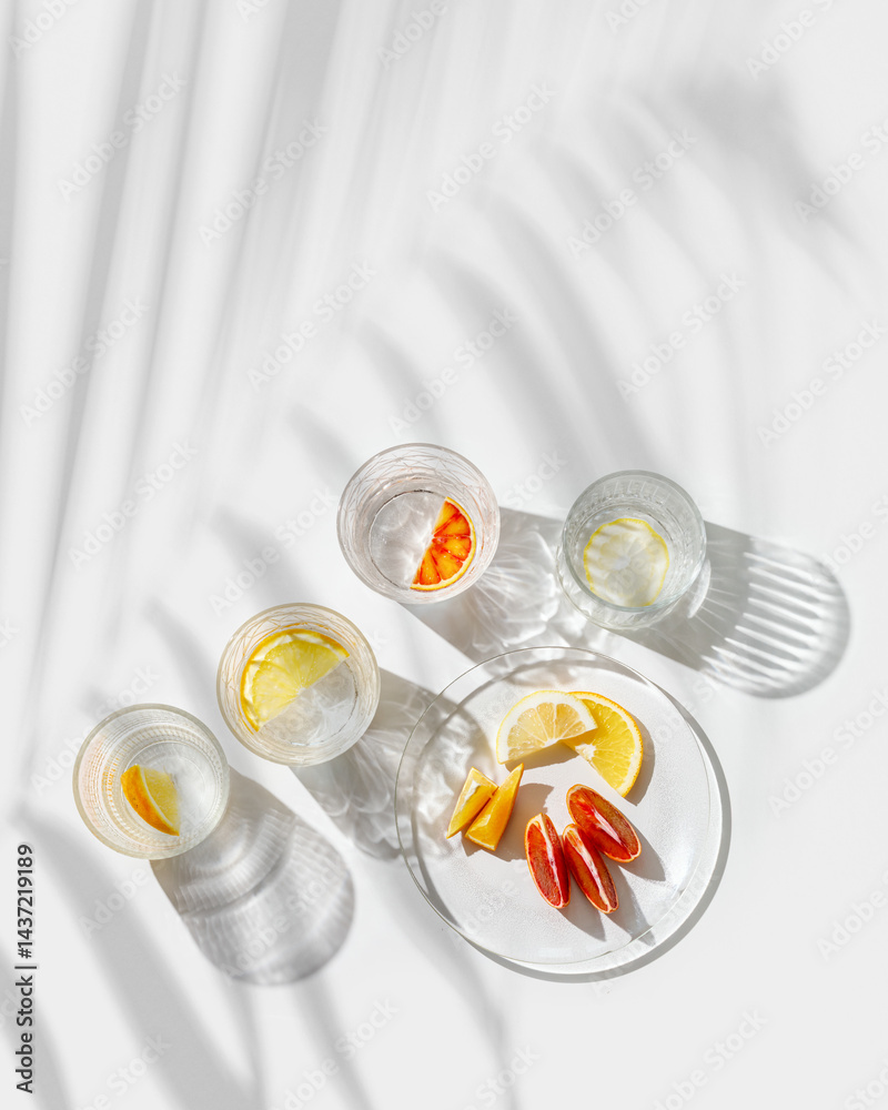 Crystal glasses with citrus slices in water on white background with palm leaf at sunlight, modern, minimalist composition with clear glass cups with water and citrus fruit, as lemon, orange