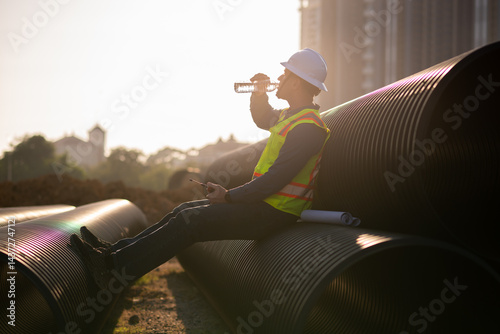 Construction worker well-deserved break, sitting large black industrial pipes site holds water bottle taking necessary breaks in physically demanding jobs.
