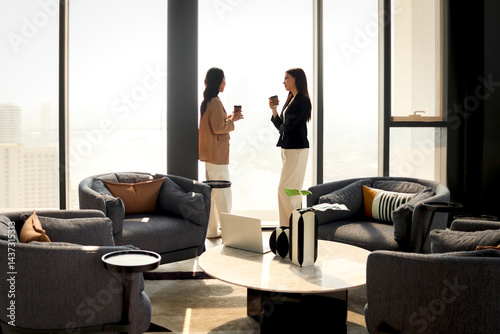 Two businesswomen have conversation during a break. Female business officers talking and drinking coffee together while standing in modern office. Businesspeople take a break at workplace.