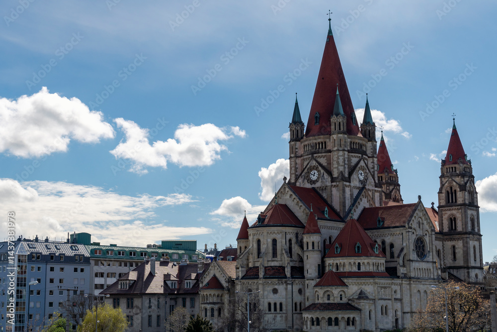 Fototapeta premium A wide-angle view of the majestic St. Francis of Assisi Church in Vienna, Austria, showcasing its distinctive red rooftops and Romanesque revival architecture against a partly cloudy blue sky.