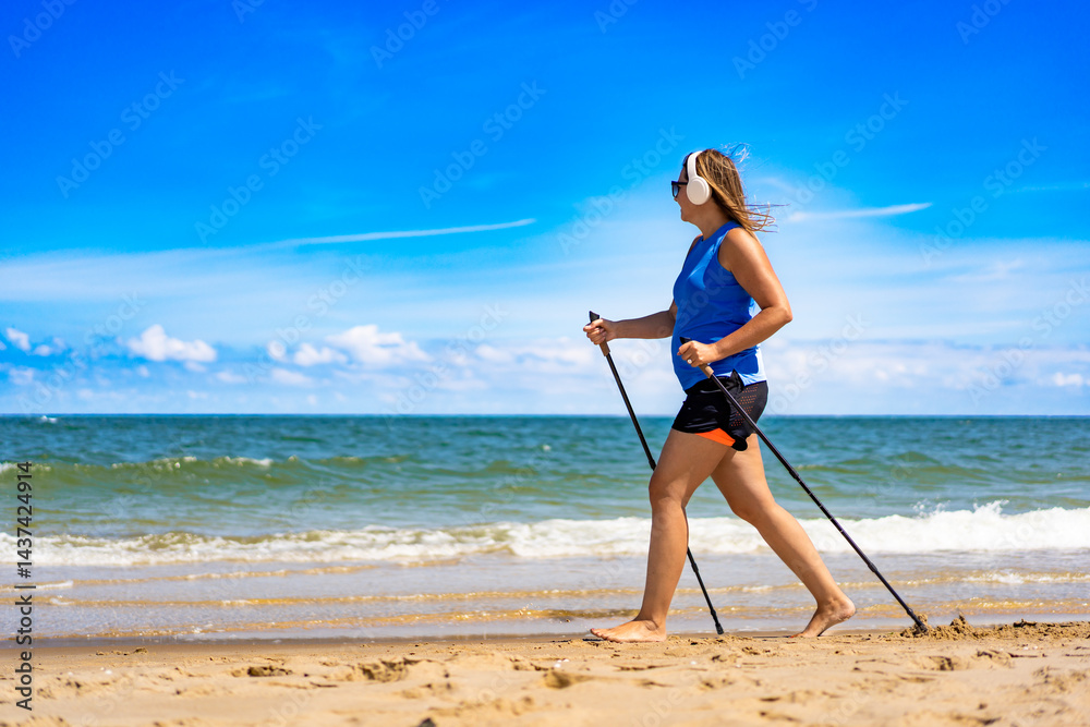 Playing sports on beach in summer. Pretty mature woman practicing Nordic walking and listening to music in headphones on sandy beach on sunny day. Side view