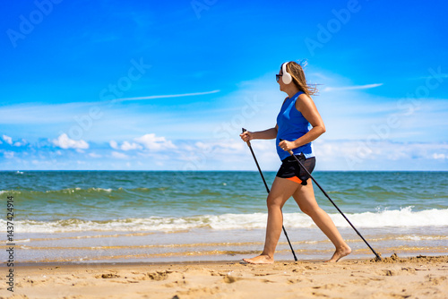 Obraz na płótnie Playing sports on beach in summer. Pretty mature woman practicing Nordic walking and listening to music in headphones on sandy beach on sunny day. Side view w salonie