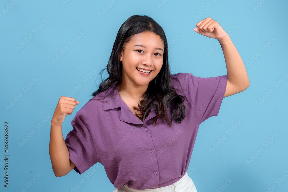 Fototapeta premium Energetic young Asian woman smiling confidently while flexing her arms in a strong pose. Her positive and enthusiastic expression is perfect for advertising, lifestyle, or promotions.