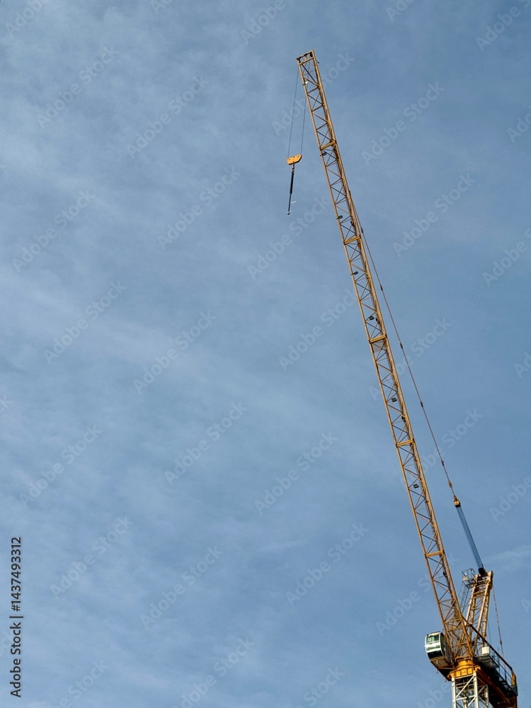 A tall yellow crane rises sharply against a soft blue sky, with its hook hanging loose, capturing the stillness and scale of an unfinished construction. A tall yellow crane rises sharply against a soft blue sky, with its hook hanging loose, capturing the stillness and scale of an unfinished construction.