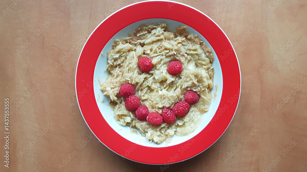 Children's plate with muesli and grated apples. A face made of raspberries. On a wooden table.