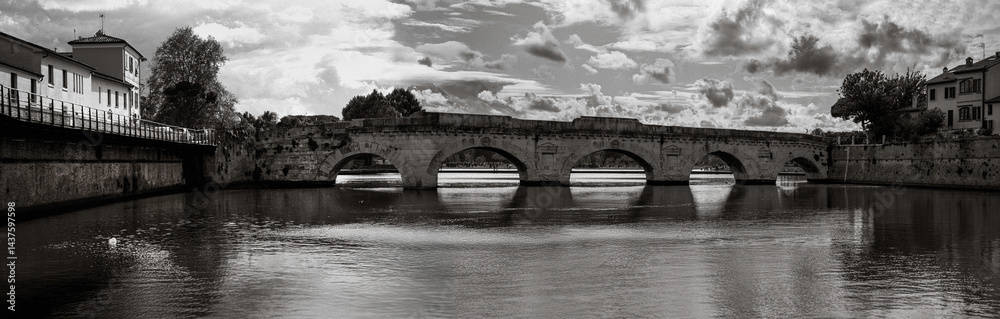 Fototapeta premium Rimini city skyline over the historic bridge Ponte di Tiberio and the Marecchia River in the Emilia-Romagna region of Italy, captured in a tranquil black and white retro-style panoramic photo