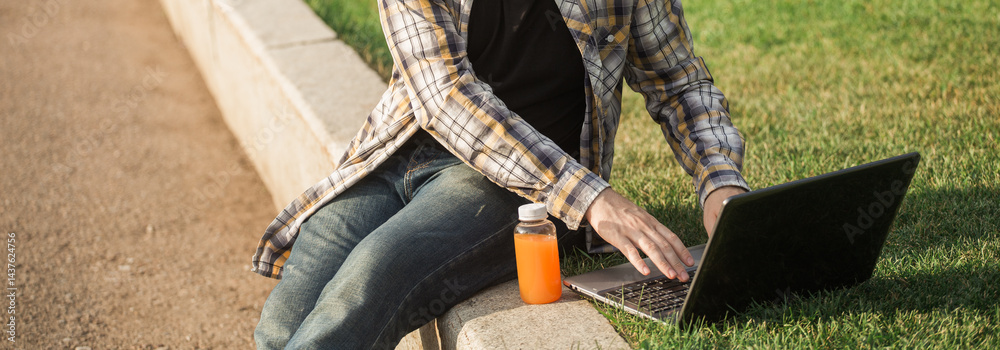 Man working outdoors on a laptop with juice beside him. Concept of remote work, productivity, and casual freelance lifestyle in urban parks. Banner with copy space
