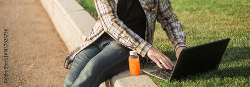 Obraz na płótnie Man working outdoors on a laptop with juice beside him. Concept of remote work, productivity, and casual freelance lifestyle in urban parks. Banner with copy space w salonie