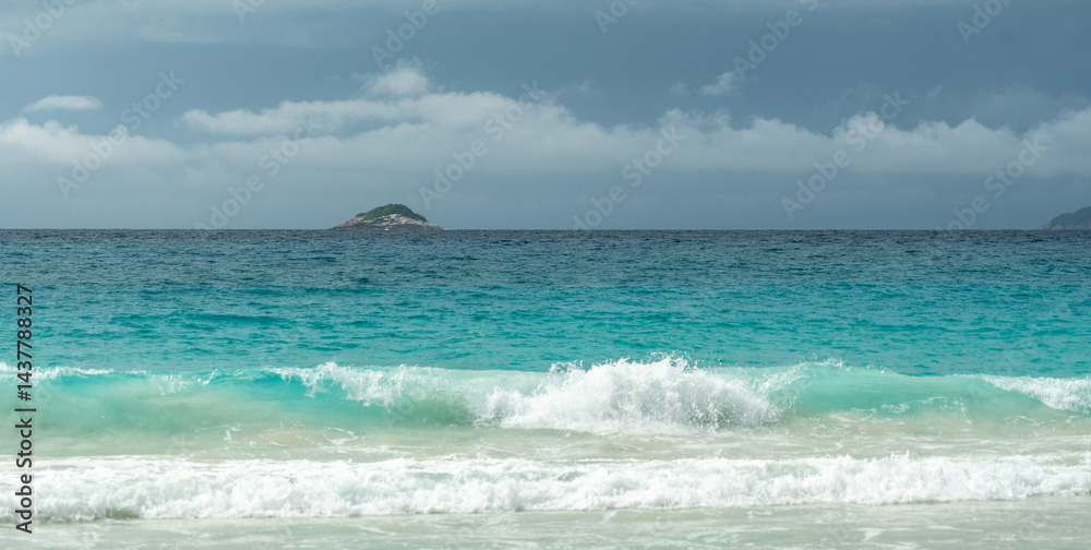 Tropical turquoise ocean waves breaking on sandy beach in Seychelles with distant island on horizon