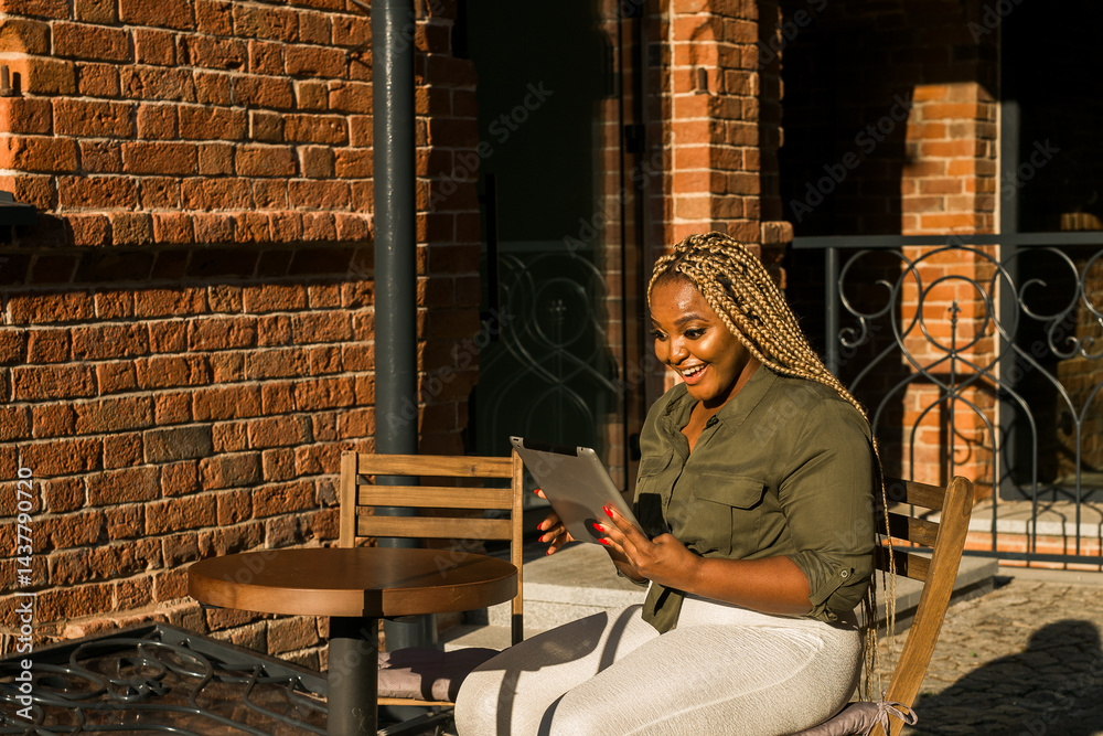 African american woman smiling and holding a tablet while sitting on outdoor cafe. Communication, happiness, and digital connection with loved ones. Banner with copy space