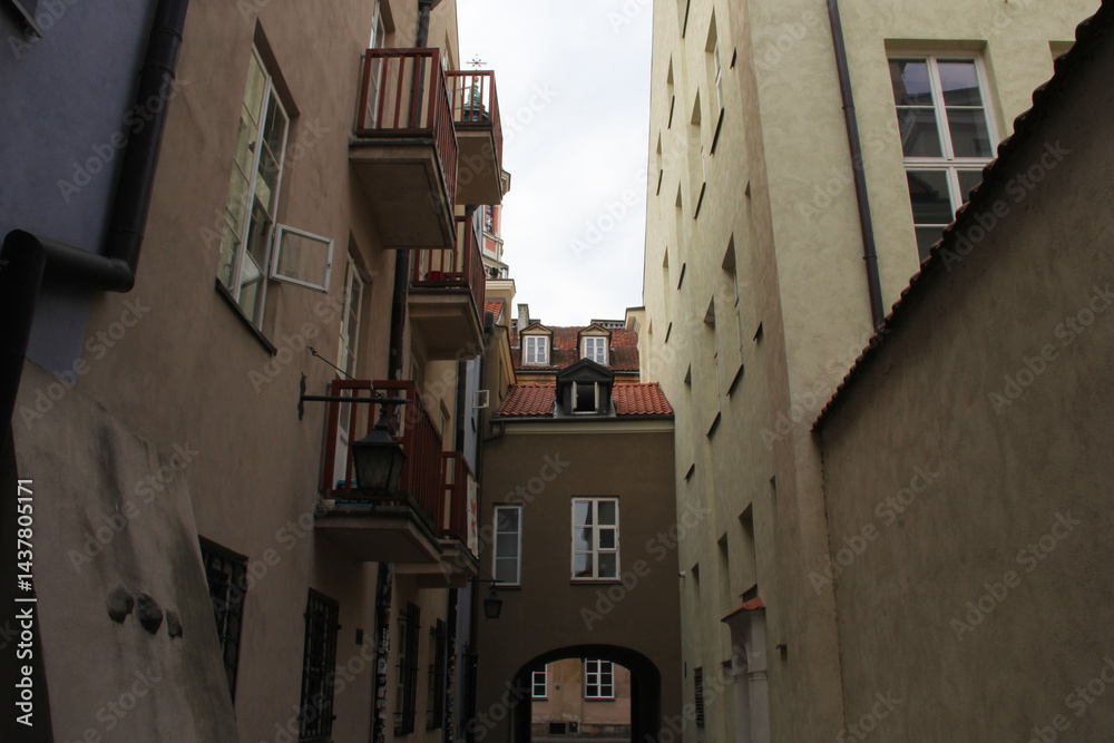Fototapeta premium Quiet narrow passageway with red balcony railings, dormer windows, and aged stucco walls in an old European city — traditional residential architecture with cozy courtyard charm