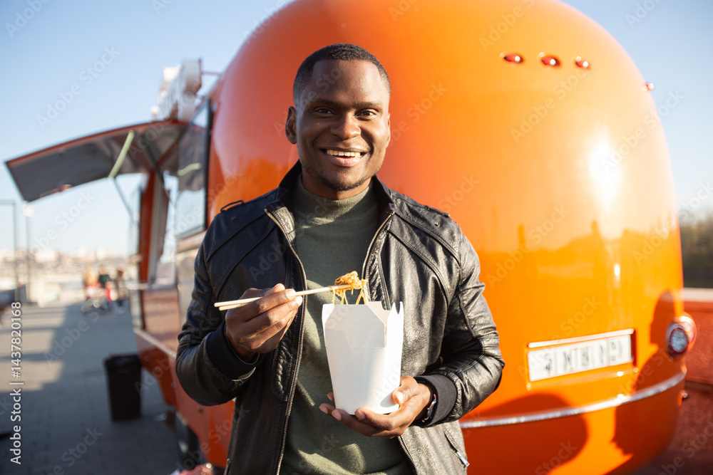 Man holding noodles and smiling on a city street near food track. Concept of urban lifestyle, street food culture and positive energy.