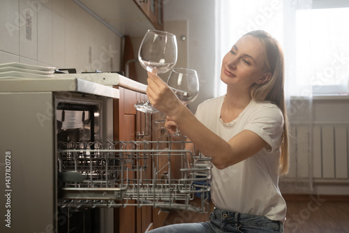 Happy woman unloading clean wine glasses from the dishwasher in a modern kitchen, enjoying her daily routine and the convenience of modern appliances