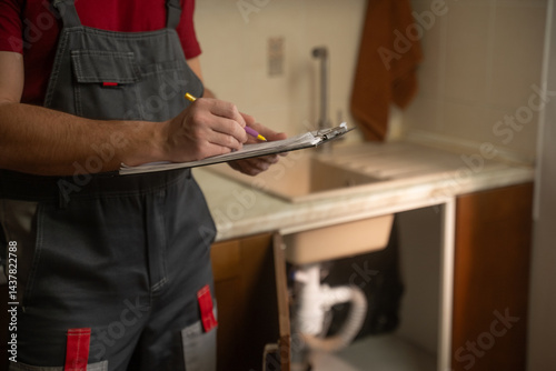 Experienced maintenance professional conducting thorough kitchen sink plumbing inspection, carefully documenting findings with clipboard and detailed notes for comprehensive service evaluation