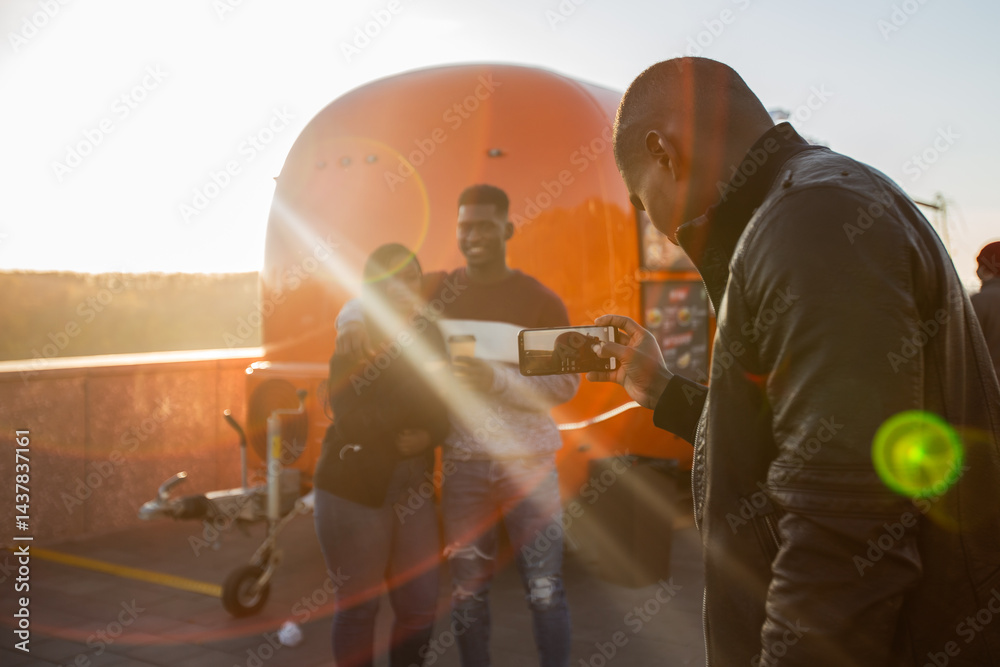 African american man photographing friends with smartphone near food truck. Concept of modern communication and leisure moments.