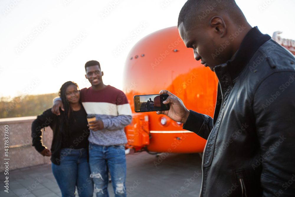 African american man photographing friends with smartphone near food truck. Concept of modern communication and leisure moments.