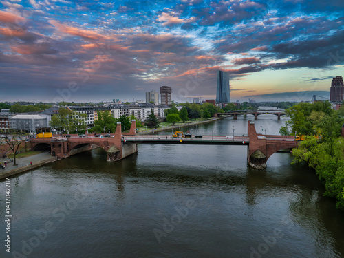 Aerial view of Frankfurt, Germany, showing the Alte Brucke over the Main River, lush greenery, and the European Central Bank at sunset.