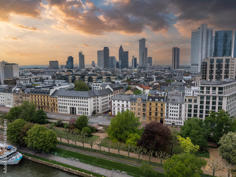 Aerial view of Frankfurt, Germany, featuring modern skyscrapers like Commerzbank Tower, a riverside promenade, a docked boat, and a dramatic sunset sky.