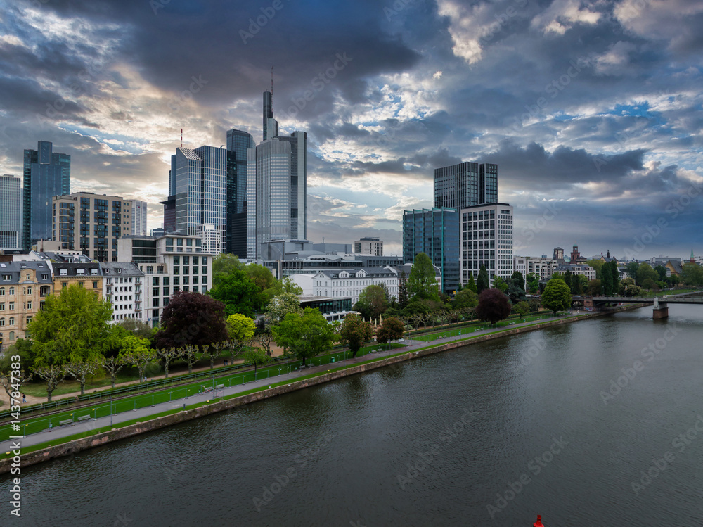 Aerial view of Frankfurt, Germany, showing modern skyscrapers like the Commerzbank Tower, the Main River, green promenades, and a cloudy sunset sky.