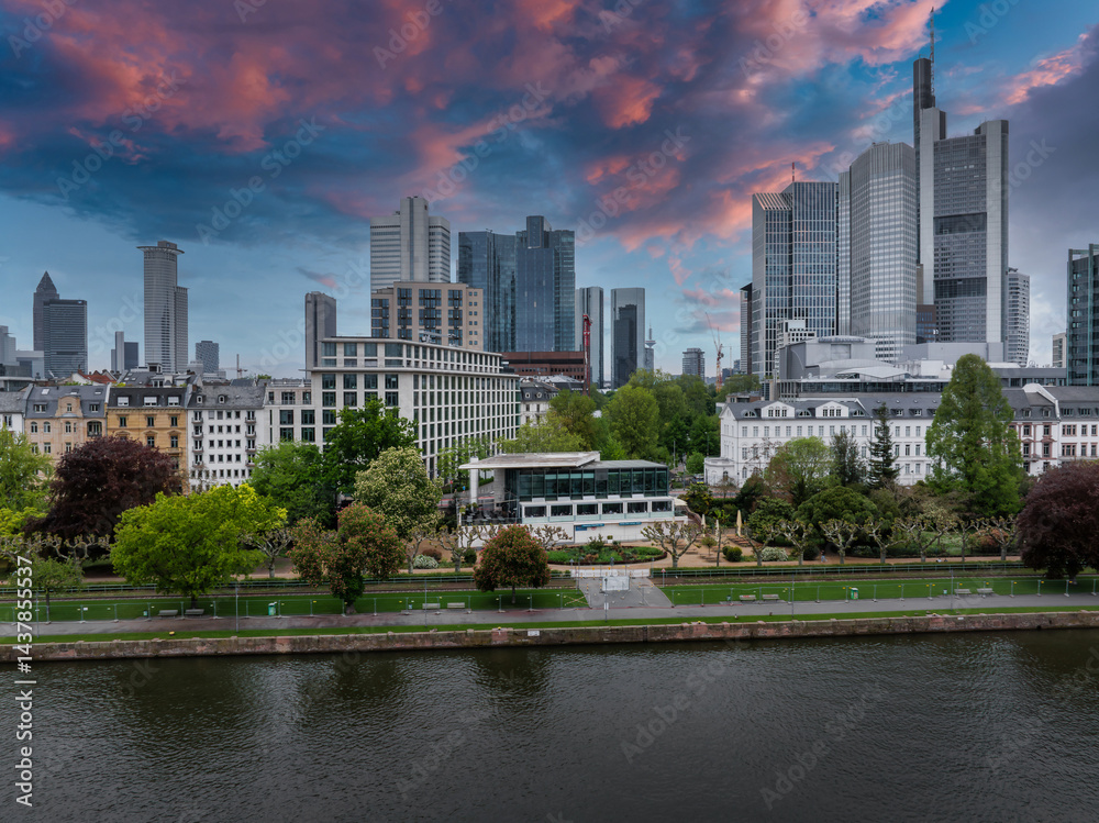Aerial view of Frankfurt, Germany, featuring the skyline with Commerzbank Tower, riverside park along the Main River, and a vibrant sunset sky.