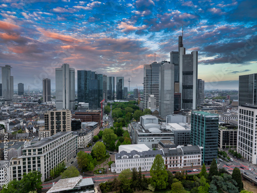 Aerial view of Frankfurt, Germany, featuring modern skyscrapers like Commerzbank Tower, historic buildings, green spaces, and a vibrant sunset sky.