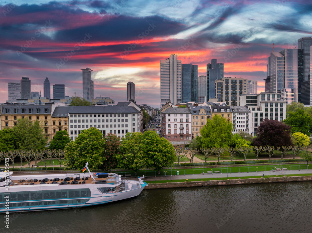 Aerial view of Frankfurt, Germany, at sunset featuring modern skyscrapers like Commerzbank Tower, the Main River, and a white riverboat by a tree lined promenade.
