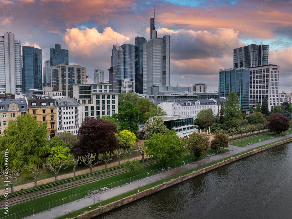 Aerial view of Frankfurt, Germany, featuring the Commerzbank Tower, riverside park greenery, and vibrant orange and pink sunset skies over the Main River.