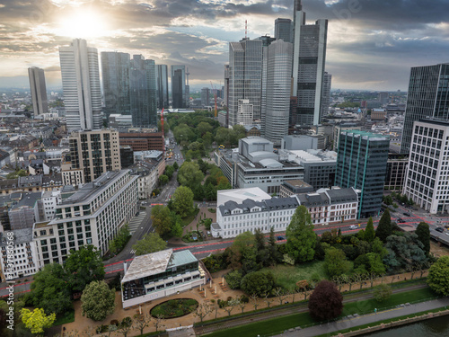 Aerial view of Frankfurt, Germany, featuring modern skyscrapers like Commerzbank Tower, a green park, the Main River, and a dramatic sunset sky.