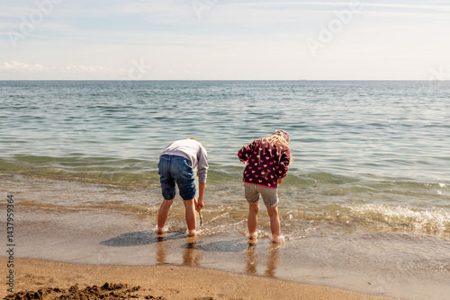 Fototapeta Naklejka Na Ścianę i Meble -  Two girls, fully dressed, playing with sand and waves and dipping their feet into the Mediterranean water, having a fun, joyful time. Children at the beach when it is too cold to swim