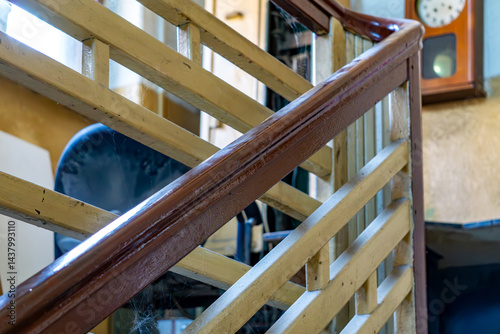 wooden balustrade of the interior stairs. The balustrade is painted yellow and the handrail is dark brown. In the background, a fragment of the interior is visible, including an old wall clock and cha