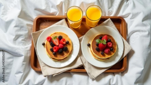 breakfast with two plates of pancakes topped with fresh berries and two glasses of orange juice on a wooden tray placed on white bed linens, concept of healthy living, hotel service, wellness retreats