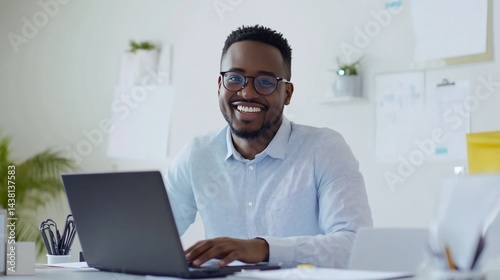 Smiling man with glasses using a laptop in a bright office workspace with plants and papers