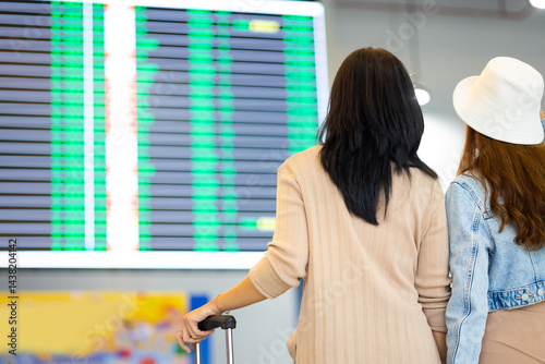 Check flight schedule on board with information display. arrival and departure board.  Two Asian woman use smartphone cheking flight for travel in long weekend