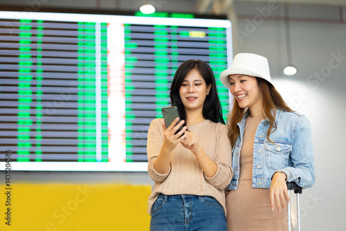 Check flight schedule on board with information display. arrival and departure board.  Two Asian woman use smartphone cheking flight for travel in long weekend