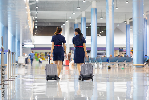 Obraz na płótnie Rear view Two woman friends walking in airport. Cabin crew, air hostess working in airplane. Service mind. Asian stewardess with suitcase at modern airport terminal. flight attendant w salonie