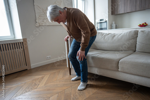 Bent senior painfully straightens legs while gripping swollen knee with degenerative joint disease. Pensioner leaning on sofa, struggle of bone erosion, synovial inflammation, cartilage loss