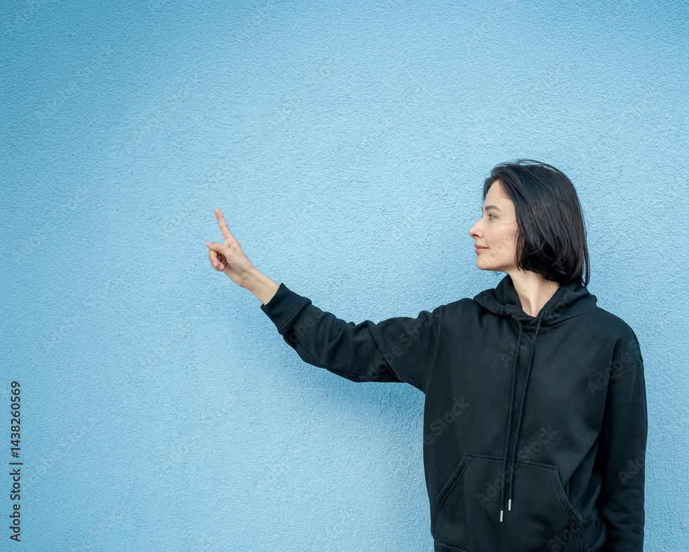 Modern girl in black sweatshirt isolated on plain bright blue background, pointing with hand to side empty space for table or chart. Photo of model on wall background with empty space for text.