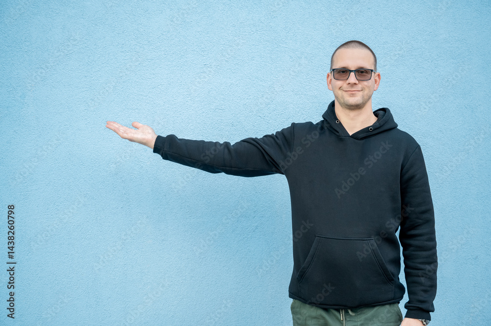 Stylish guy in glasses and black sweatshirt isolated on plain bright blue background, pointing with hand to side empty space for table or graph. Photo of model on wall background with empty space