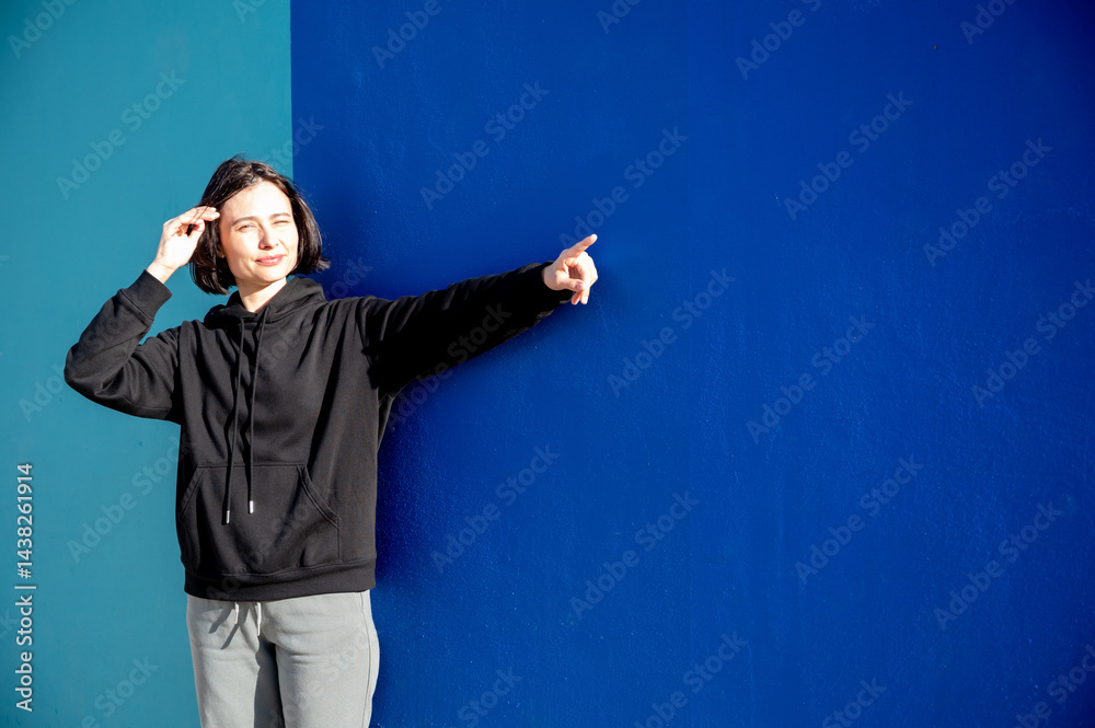 Modern girl in black sweatshirt isolated on plain bright blue background, pointing with hand to side empty space for table or chart. Photo of model on wall background with empty space for text.