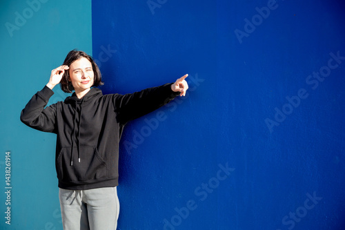 Obraz na płótnie Modern girl in black sweatshirt isolated on plain bright blue background, pointing with hand to side empty space for table or chart. Photo of model on wall background with empty space for text. w salonie