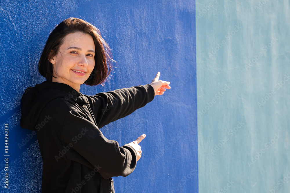 Modern girl in black sweatshirt isolated on plain bright blue background, pointing with hand to side empty space for table or chart. Photo of model on wall background with empty space for text.