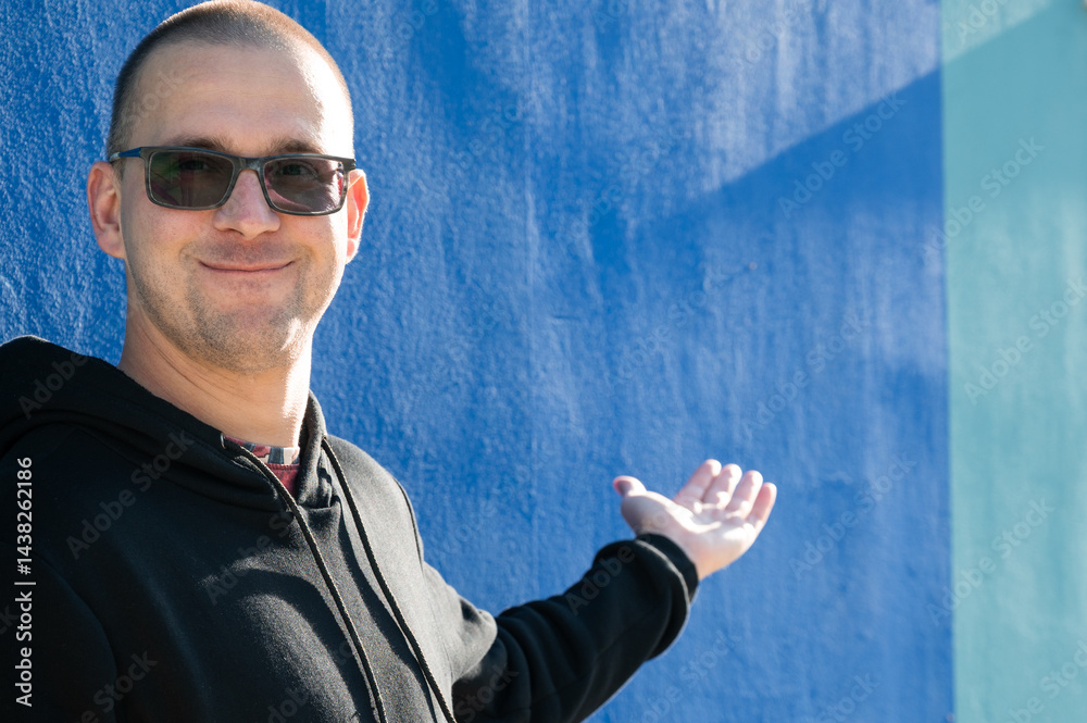 Stylish guy in glasses and black sweatshirt isolated on plain bright blue background, pointing with hand to side empty space for table or graph. Photo of model on wall background with empty space