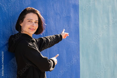 Obraz na płótnie Modern girl in black sweatshirt isolated on plain bright blue background, pointing with hand to side empty space for table or chart. Photo of model on wall background with empty space for text. w salonie
